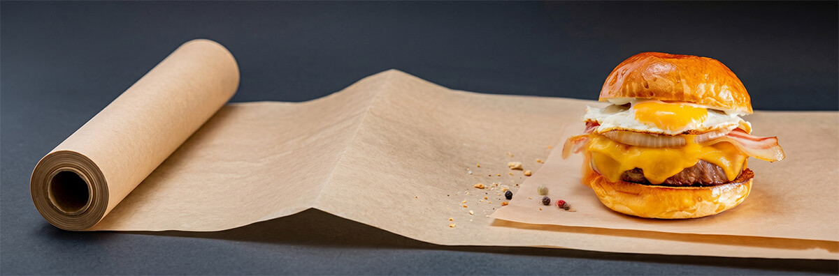 Unbleached PFAS-free greaseproof paper roll unrolled beneath a cheeseburger, demonstrating oil resistance